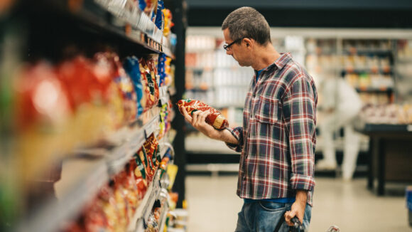 Man standing in a supermarket aisle and reading the label on a bag of pasta while grocery shopping. Image depicts consumer insight.