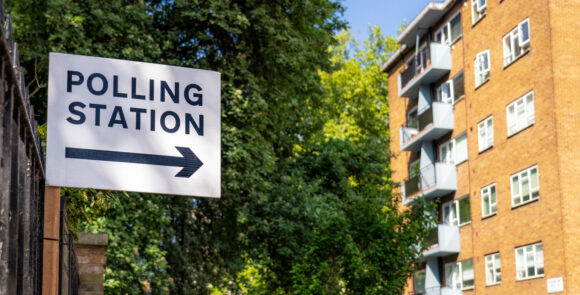 Image depicts a polling station sign and a block of flats in the UK