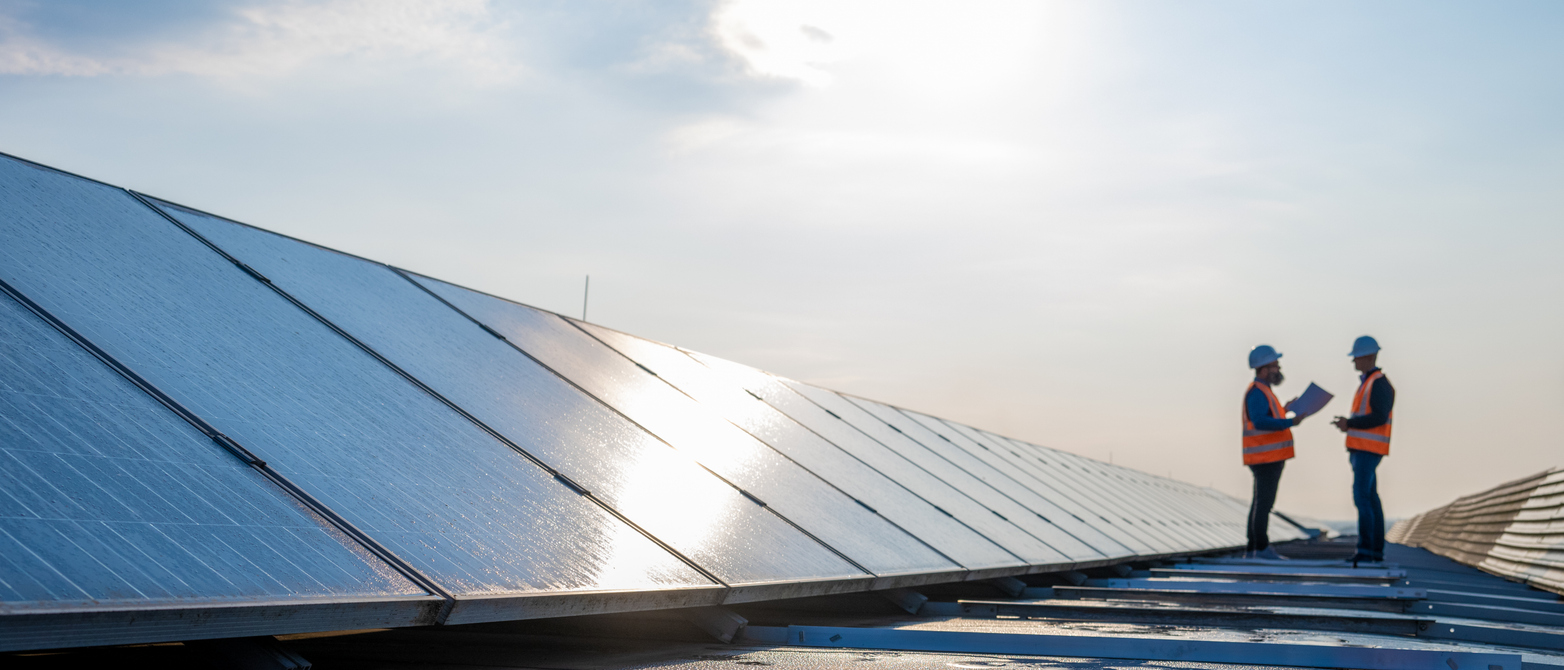 Two men stand on a roof between rows of net zero photovoltaic panels